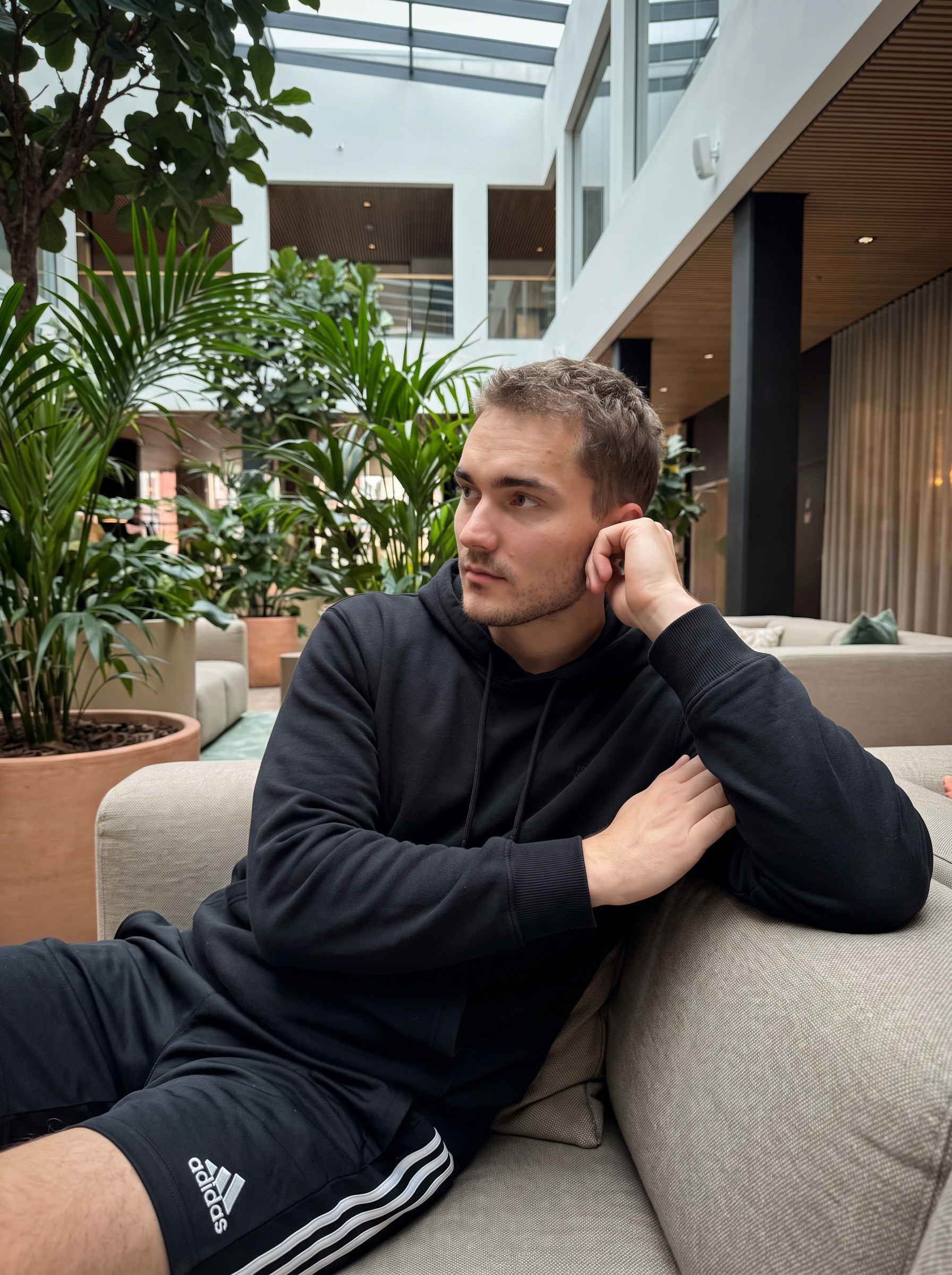 Subject: [Blank]

An ultra-detailed, HD, authentic portrait captured as if taken by an iPhone camera, subject seated in a relaxed, slumped posture, body angled slightly right, head turned sharply left, looking off-camera. Right arm is bent, hand resting on the side of the face near the ear. Left arm is bent, elbow resting on the sofa arm, forearm across the body. Legs are bent and partially visible, implying a relaxed, drawn-up position. Outfit: loose-fitting black hoodie with visible fabric creases, and black athletic shorts featuring two horizontal white stripes on the side seams. Background: lush indoor environment with abundant green foliage and large potted plants, suggesting a hotel lobby or a similar lounge area, with subtle hints of modern architecture and ambient lighting in the distant background. Mid-shot, soft ambient lighting from overhead and background sources, creating a natural, slightly dim appearance with moderate contrast. shot on iPhone (Apple ProRAW / RAW-like capture, 12-bit DNG look), handheld smartphone capture, mild wide-angle (~24–28mm equivalent), Smart HDR + Deep Fusion behavior, natural micro-contrast, neutral auto white balance, realistic exposure roll-off, tiny sensor noise, fine grain in shadows, subtle lens distortion and mild vignetting, phone-like depth behavior (not DSLR bokeh), no beauty filter, no skin smoothing. Avoid AI artifacts: no waxy or plastic textures, no over-sharpening halos, no CGI surfaces, no warped anatomy, no extra fingers, no distorted hands, no fake depth blur. the body pose is an exact 1:1 clone of the reference image, with identical posture, limb positioning, weight distribution, and head tilt, only the subject’s appearance is neutralized while preserving the physical pose perfectly. no color grading, no stylization, no cinematic look, and the tone must be neutral (not too cool, not too warm). Include subtle iPhone realism cues (mild wide-angle perspective, ~24–28mm equivalent, f/1.9, 1/125s, ISO 125), tiny sensor noise, and natural exposure roll-off; portrait orientation, aspect ratio 4:3, and it must not look AI-generated—just like a real, imperfect iPhone snapshot. | User Input: N/A