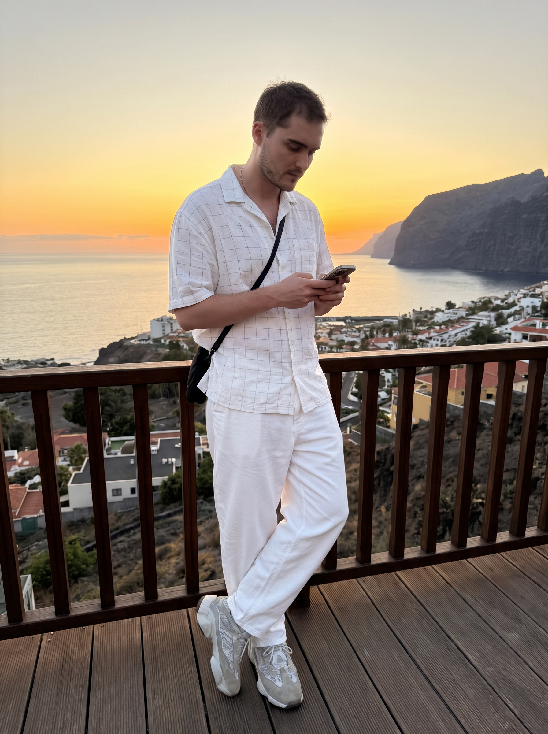 Subject: [Blank]
An ultra-detailed, HD, authentic portrait captured as if taken by an iPhone camera, torso angled slightly left, head tilted down looking at a smartphone held in both hands in front of the waist. Relaxed standing posture, legs slightly apart with the left foot positioned slightly forward. Outfit: loose-fitting white short-sleeved button-down shirt with a subtle grid texture, unbuttoned at the collar, baggy white trousers with natural wrinkles and folds, a thin black cross-body bag strap resting across the chest, and grey and white chunky suede sneakers. Background: outdoor wooden balcony deck with a dark railing, overlooking a coastal sunset. Vibrant orange and yellow sunset sky over calm ocean water, dark rugged cliffs to the right, and a coastal town with a winding road below. Full-body shot, soft ambient sunset lighting with the subject in natural, slightly shadowed evening light. shot on iPhone (Apple ProRAW / RAW-like capture, 12-bit DNG look), handheld smartphone capture, mild wide-angle (~24–28mm equivalent), Smart HDR + Deep Fusion behavior, natural micro-contrast, neutral auto white balance, realistic exposure roll-off, tiny sensor noise, fine grain in shadows, subtle lens distortion and mild vignetting, phone-like depth behavior (not DSLR bokeh), no beauty filter, no skin smoothing. Avoid AI artifacts: no waxy or plastic textures, no over-sharpening halos, no CGI surfaces, no warped anatomy, no extra fingers, no distorted hands, no fake depth blur. the body pose is an exact 1:1 clone of the reference image, with identical posture, limb positioning, weight distribution, and head tilt, only the subject’s appearance is neutralized while preserving the physical pose perfectly. Do not change Subject's facial features. Subjects must look 1000% identical to uploaded images. no color grading, no stylization, no cinematic look, and the tone must be neutral (not too cool, not too warm). Include subtle iPhone realism cues (mild wide-angle perspective, ~24–28mm equivalent, f/1.9, 1/125s, ISO 125), tiny sensor noise, and natural exposure roll-off; portrait orientation, aspect ratio 3:4, and it must not look AI-generated—just like a real, imperfect iPhone snapshot. | User Input: N/A