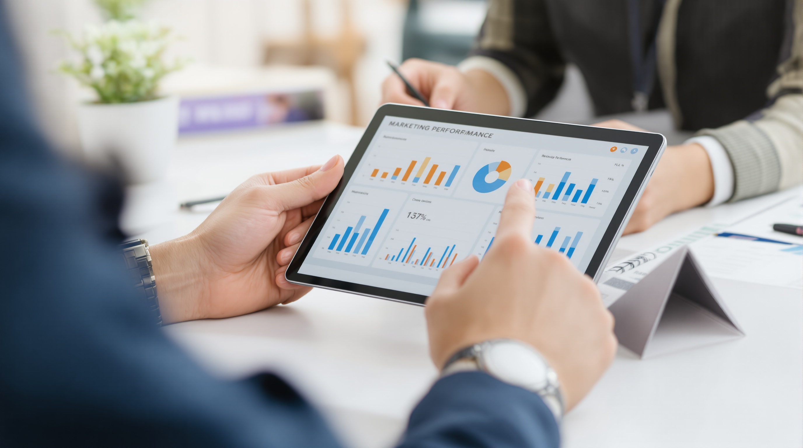 Close-up of two pairs of hands analyzing marketing performance charts on a digital tablet during a team meeting, with a blurred office background.
