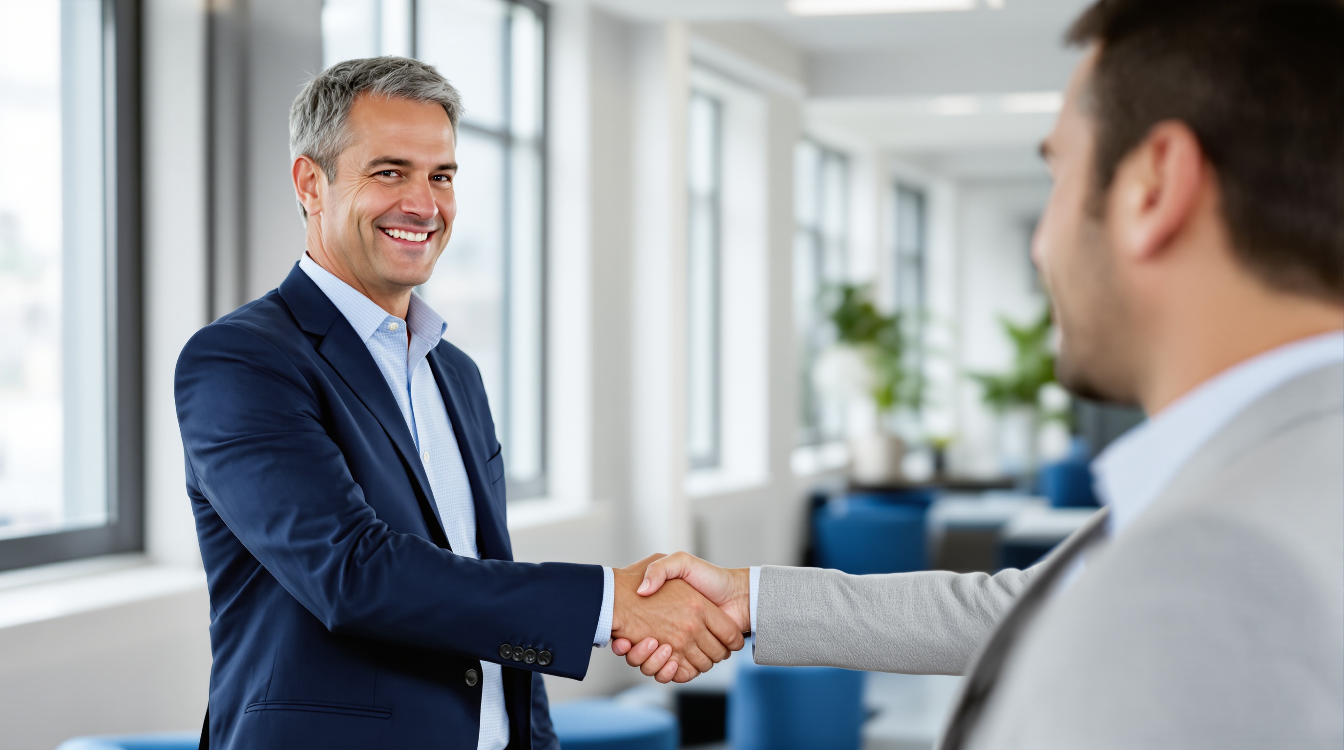 A confident executive in a navy suit shaking hands with a consultant in a bright, modern office, symbolizing partnership and collaboration.